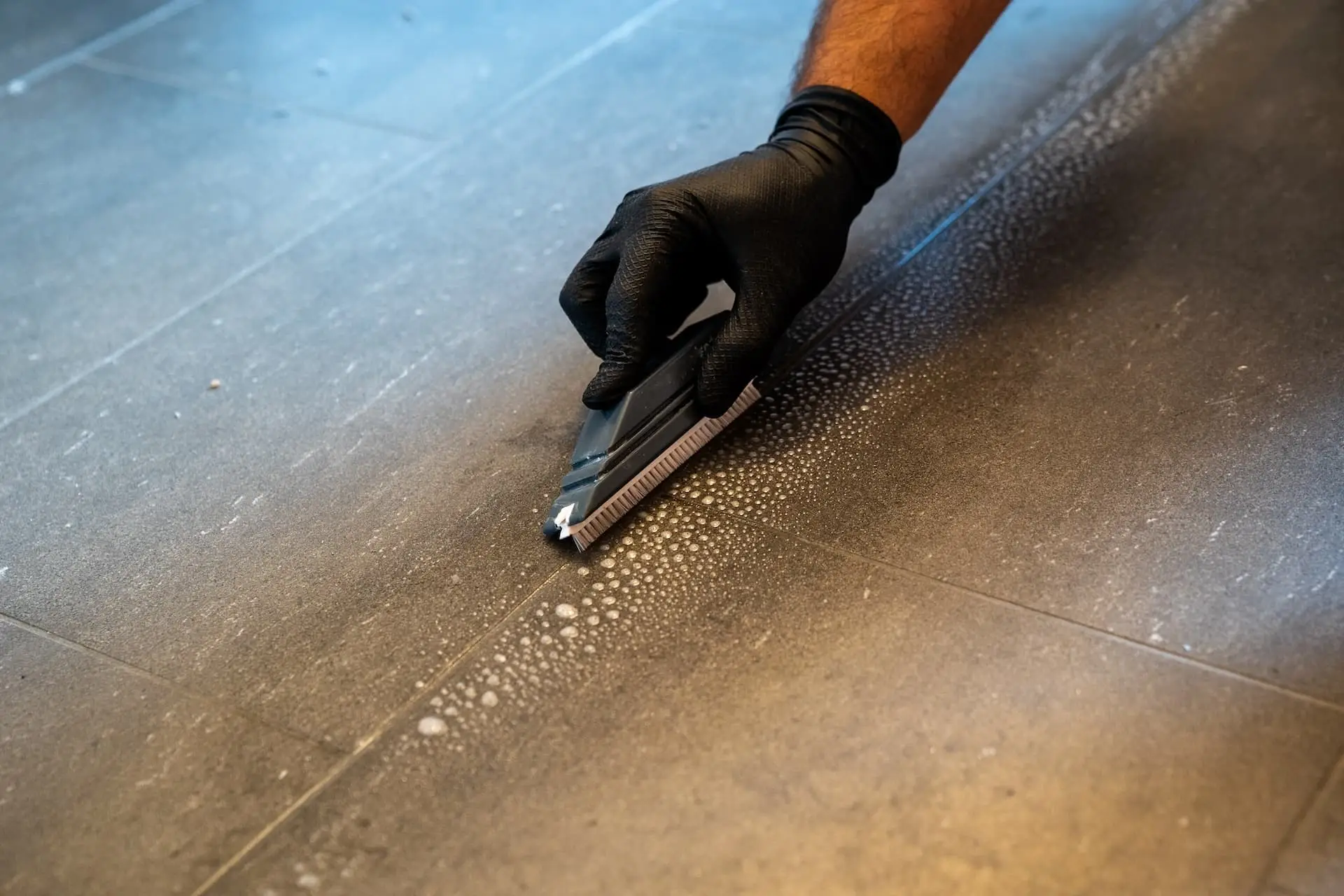 A gloved hand using a scrub brush to clean a tile floor, revealing a pattern of debris