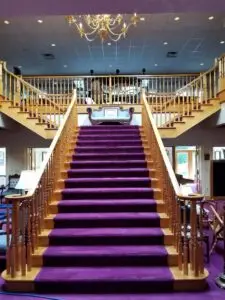 A grand staircase with a plush purple carpet leading up to an ornate chandelier and a second level balcony. The stairs and railings are made of polished wood.