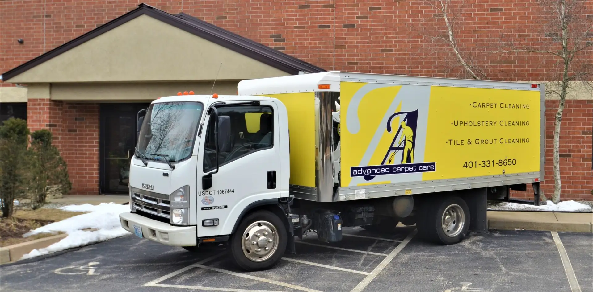 A white commercial truck with the company name 'Advanced Carpet Care' and the services 'Carpet Cleaning, Upholstery Cleaning, Tile & Grout Cleaning' printed on the side is parked in a parking lot in front of a brick building.