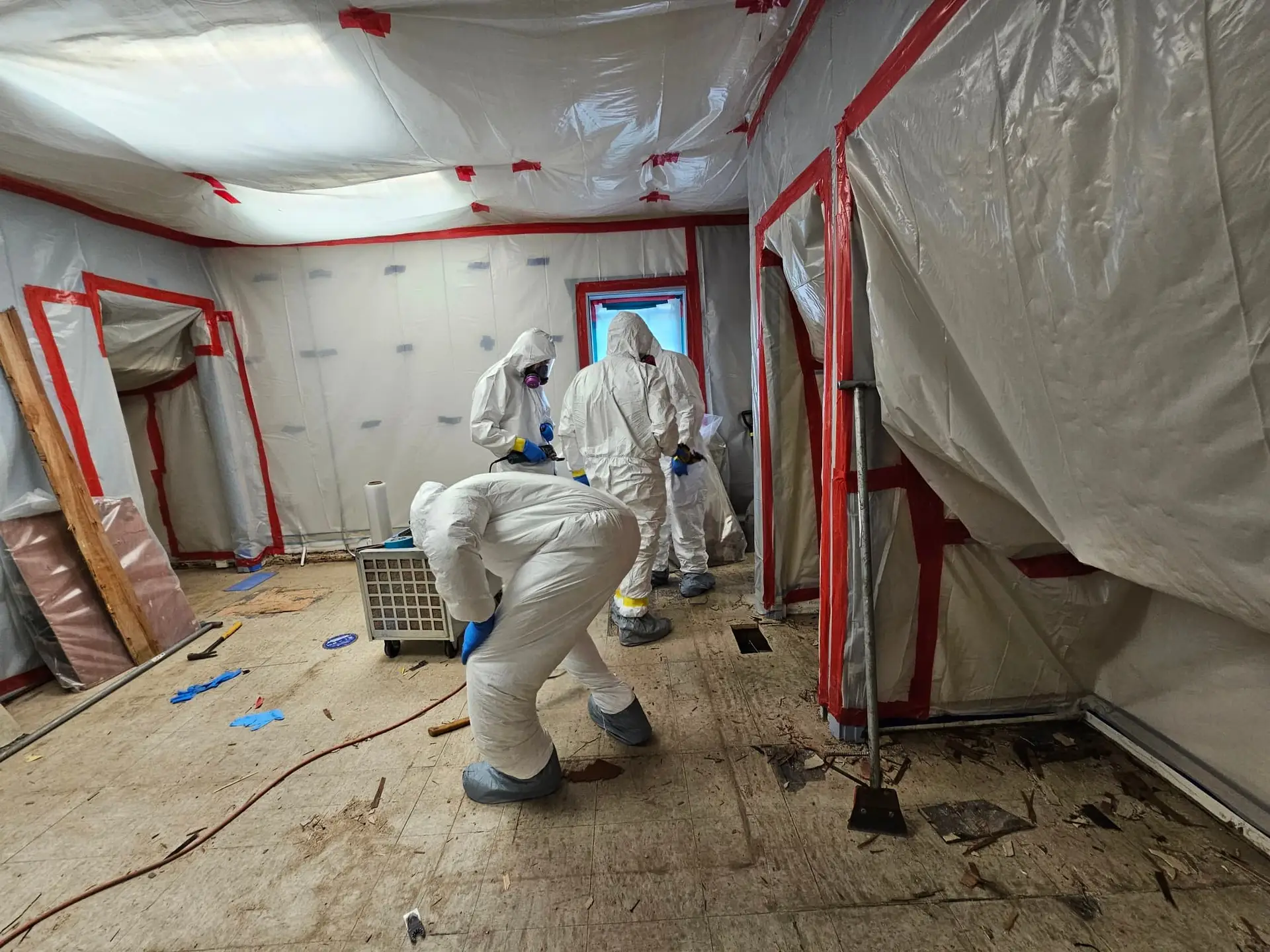 The image shows two people in white hazmat suits working in a room surrounded by plastic sheeting.