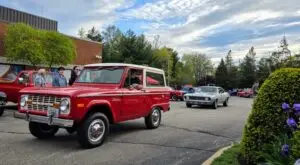 A red vintage Ford Bronco SUV parked in a parking lot, surrounded by other classic cars. The image captures the details and design of the Bronco, including its distinctive shape and large tires.