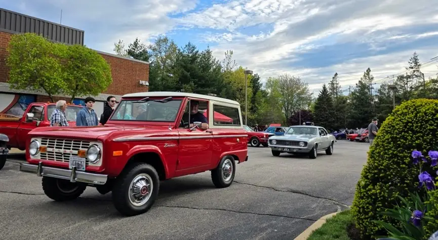 A red vintage Ford Bronco SUV parked in a parking lot, surrounded by other classic cars. The image captures the details and design of the Bronco, including its distinctive shape and large tires.