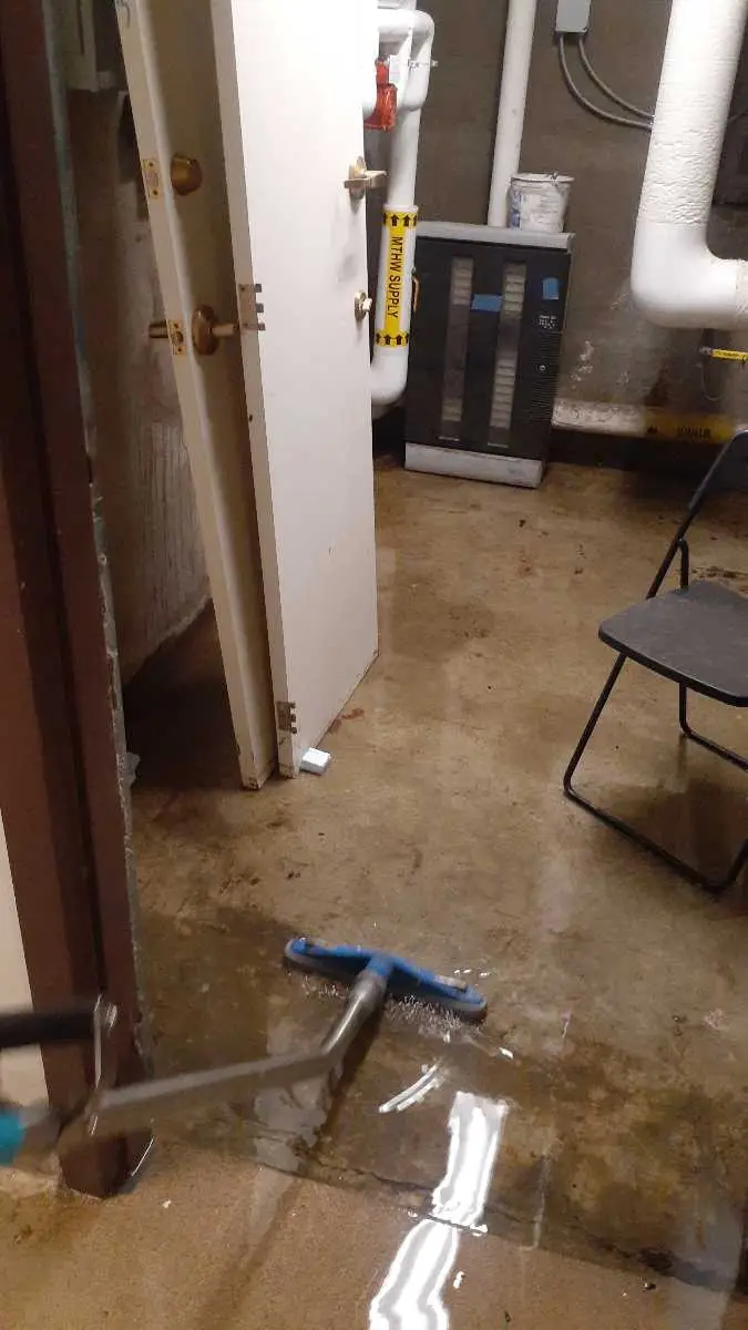 A flooded basement with water on the floor, a black electrical panel, and various pipes and equipment visible.