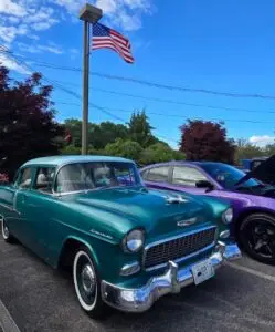 A classic American car, a vintage green Chevrolet, parked on a street with a bright blue sky and trees in the background, with a flag pole flying an American flag.