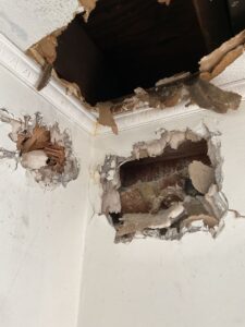 An image showing extensive damage and holes in a wall, with exposed wooden framing and crumbling drywall or plaster.