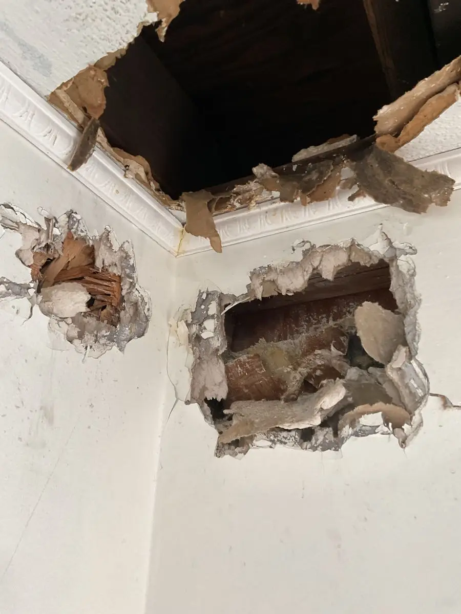 An image showing extensive damage and holes in a wall, with exposed wooden framing and crumbling drywall or plaster.