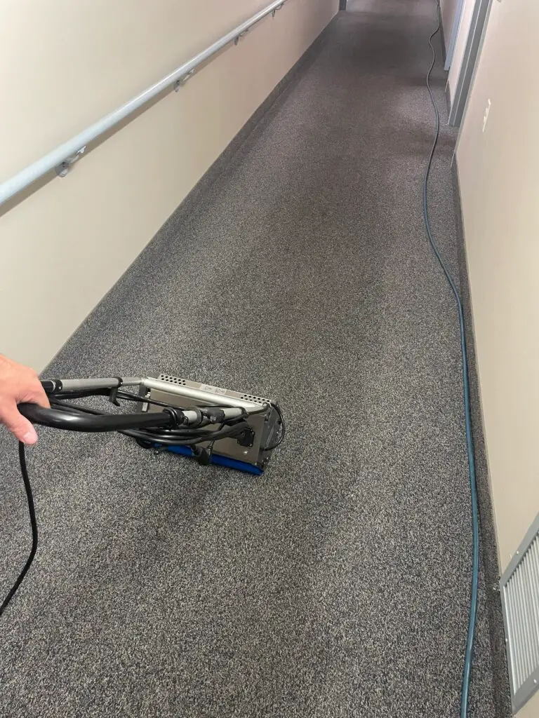 A view of a carpeted hallway, with a vacuum cleaner in the foreground being used to clean the floor.
