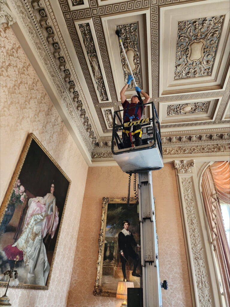 Technician on a lift carefully cleaning an ornate, decorative ceiling inside a historic room with detailed moldings, large framed portraits, and elegant interior finishes.
