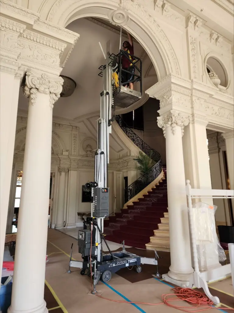 Technician standing on a lift cleaning ornate architectural detailing high above the grand entrance of a historic building, featuring large columns, intricate molding, and a sweeping staircase with a red carpet.